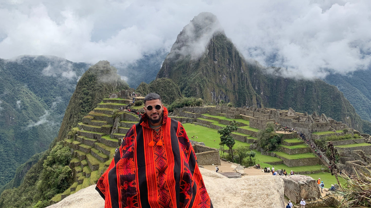 A man wearing a red and black patterned knit poncho sits on a stone ledge at Machu Picchu, with the ancient ruins, green terraces, and misty mountain peaks in the background.