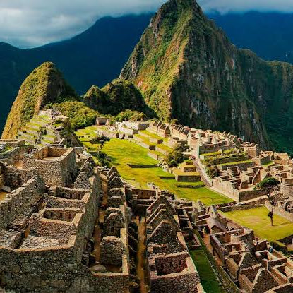 A scenic aerial view of Machu Picchu with ancient stone ruins, green terraces, and steep mountains in the background under a cloudy sky.