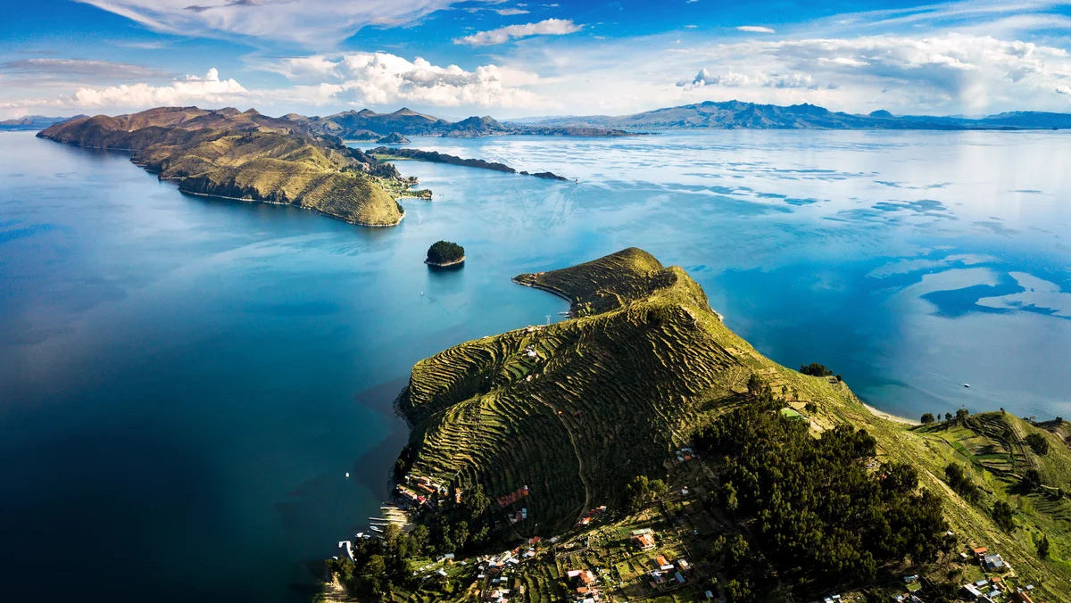 Aerial view of a dramatic mountainous lake landscape with steep green ridges, terraced fields, distant islands, and a bright blue sky with scattered white clouds.