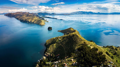 Aerial view of a dramatic mountainous lake landscape with steep green ridges, terraced fields, distant islands, and a bright blue sky with scattered white clouds.