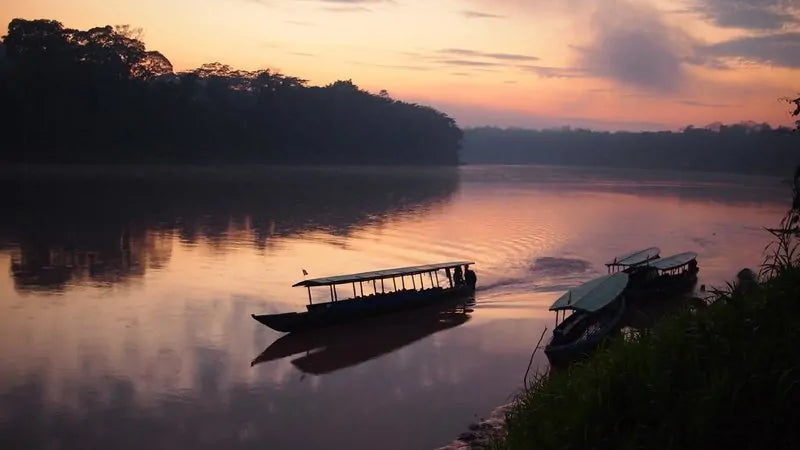 A scenic sunset over a calm river with silhouetted boats on the water and dark tree-lined banks in the background.